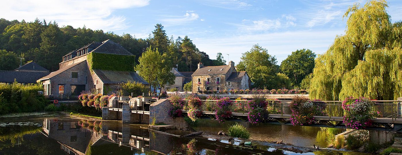 Country estate with buildings and flowers reflected in calm water, under a blue sky.